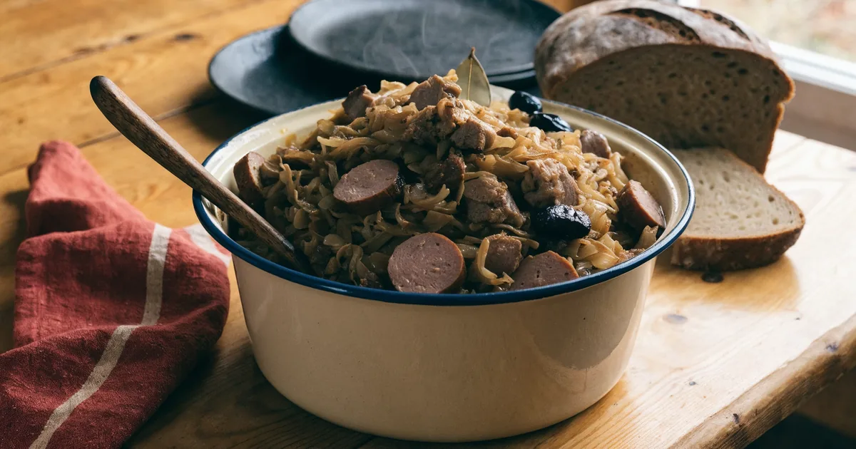Bigos z kiszonej kapusty w emaliowanym garnku, z kiełbasą, wieprzowiną i suszonymi śliwkami, obok chleb