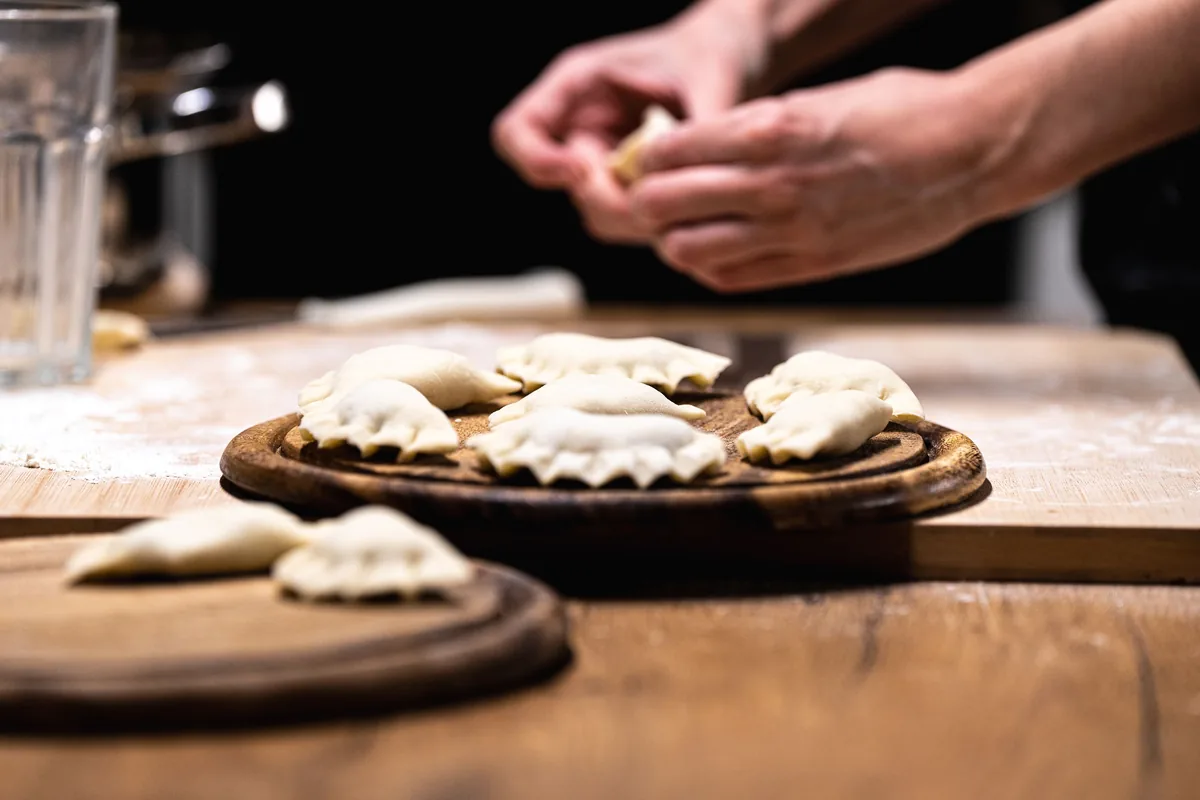 Dłonie sklejające brzeg pieroga, obok ulepione pierogi na drewnianej desce